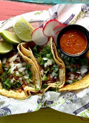 Three street tacos filled with chopped meat, onions, and cilantro, served with lime wedges, sliced radishes, and a small cup of red salsa on branded paper in a basket on a colorful wooden table.