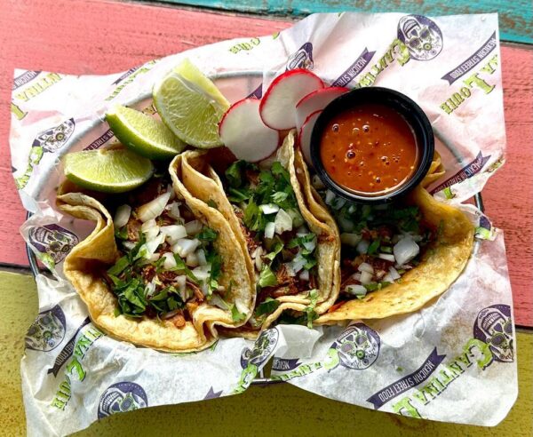 Three street tacos filled with chopped meat, onions, and cilantro, served with lime wedges, sliced radishes, and a small cup of red salsa on branded paper in a basket on a colorful wooden table.