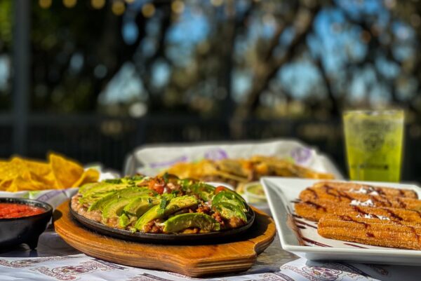 A close-up of a table filled with Mexican food, featuring a sizzling dish topped with avocado, churros on a white plate, tortilla chips with salsa, and a drink, all set outdoors with trees in the background.