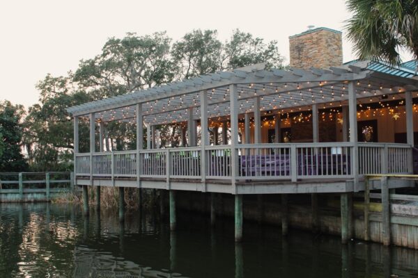 A wooden patio with string lights extends over calm water, surrounded by trees. The covered deck is attached to a building with a stone chimney and offers a peaceful waterfront view.