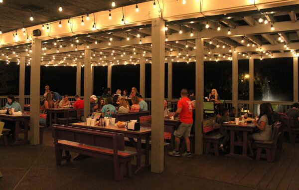 People dine and socialize at picnic tables under string lights on a covered outdoor patio at night. The atmosphere is lively and warm, with groups eating, talking, and enjoying the evening.