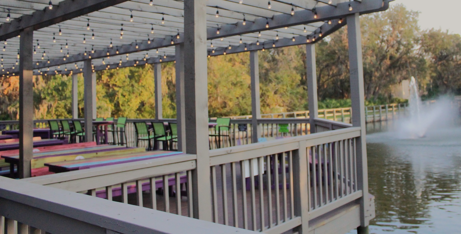 A lakeside outdoor patio with picnic tables and string lights overhead, surrounded by a wooden railing. Trees and a small fountain in the water are visible in the background.