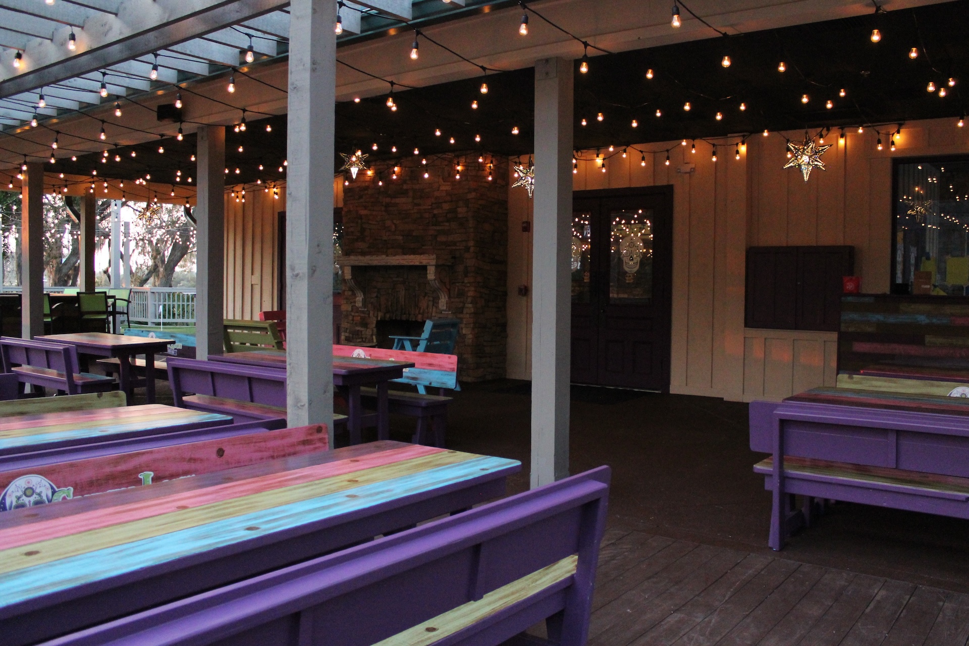 Outdoor covered patio with colorful wooden benches and tables, string lights hanging from the ceiling, and a stone fireplace in the background, creating a cozy, inviting atmosphere.