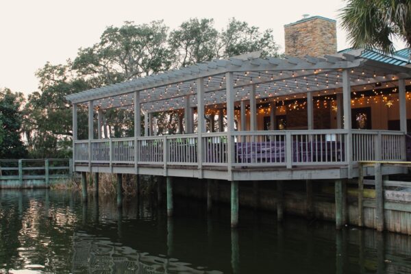 A wooden deck with string lights and a stone chimney overlooks a calm body of water, surrounded by trees and greenery in the background.