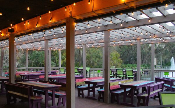 Covered outdoor patio with strings of lights overhead, featuring colorful wooden picnic tables and benches. Trees and greenery surround the space, and a water fountain is visible in the background.