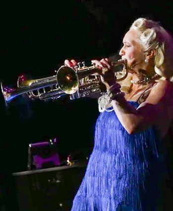 A woman in a blue fringed dress plays a trumpet on stage under dramatic lighting, with her blonde hair styled in loose waves.
