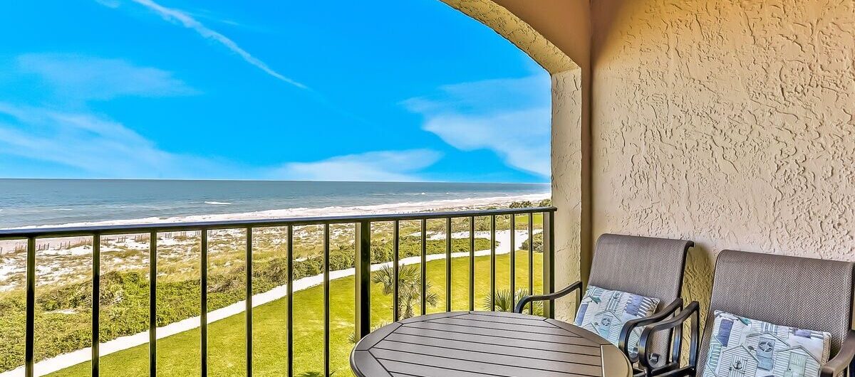 Two cushioned chairs and a round table sit on a covered balcony with a black railing, overlooking a grassy area and a sandy beach with ocean waves under a bright blue sky.