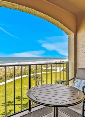 Two cushioned chairs and a round table sit on a covered balcony with a black railing, overlooking a grassy area and a sandy beach with ocean waves under a bright blue sky.