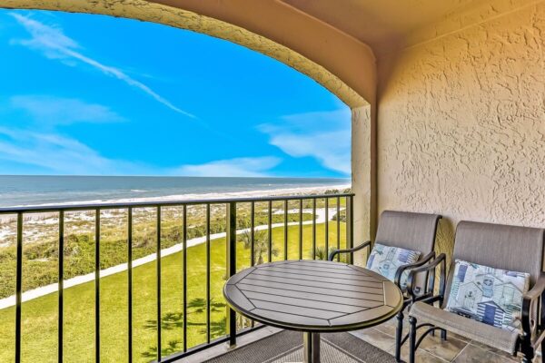 Two cushioned chairs and a round table sit on a covered balcony with a black railing, overlooking a grassy area and a sandy beach with ocean waves under a bright blue sky.