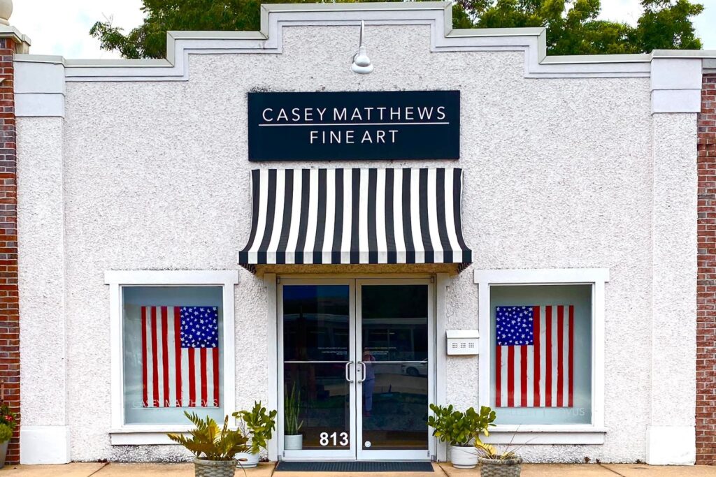 A white building with a black-and-white striped awning, labeled Casey Matthews Fine Art. Two large American flags hang in the front windows, and potted plants sit by the entrance. The address 813 is visible on the door.