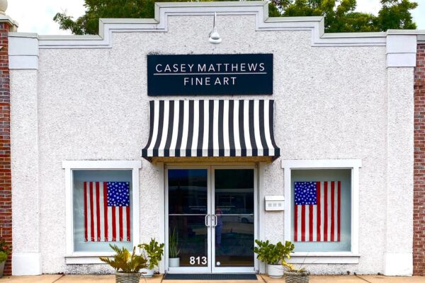 A white building with a black-and-white striped awning, labeled Casey Matthews Fine Art. Two large American flags hang in the front windows, and potted plants sit by the entrance. The address 813 is visible on the door.