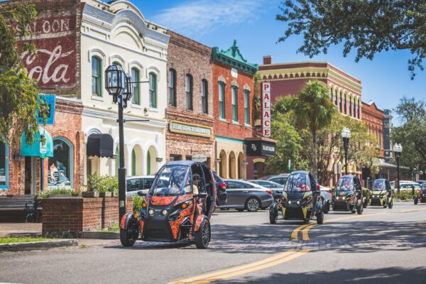 Colorful, three-wheeled electric vehicles drive down a vibrant downtown street lined with historic brick buildings, including a “Palace” theater and a faded Coca-Cola mural, on a sunny day.
