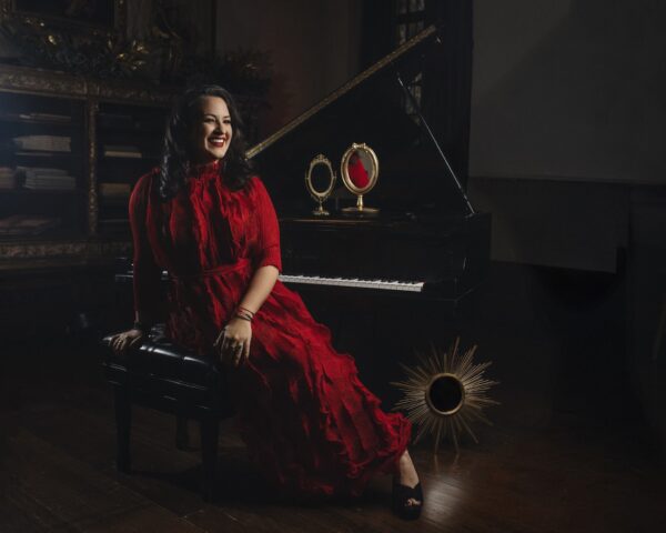 A woman in a flowing red dress sits on a piano bench, smiling. Behind her is a grand piano with a decorative mirror and sunburst ornament. The setting is elegant and warmly lit.