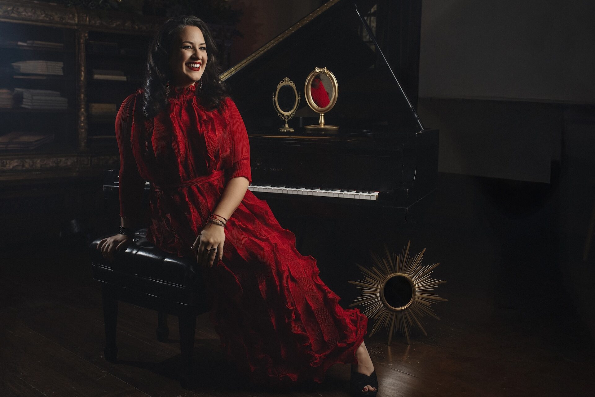 A woman in a flowing red dress sits on a piano bench, smiling. Behind her is a grand piano with a decorative mirror and sunburst ornament. The setting is elegant and warmly lit.