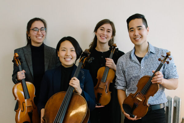 Four young adults stand smiling, each holding a string instrument—two violins, a viola, and a cello—against a plain light-colored background.