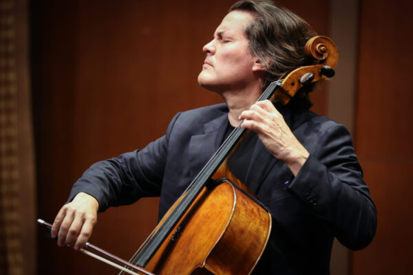 A man with long hair closed eyes passionately plays a cello on stage, wearing a dark suit, against a blurred brown background.