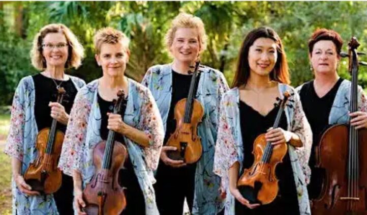 Five women stand outdoors, each holding a string instrument—violins, viola, and cello. They wear matching floral-patterned jackets over black outfits and smile at the camera, with greenery in the background.