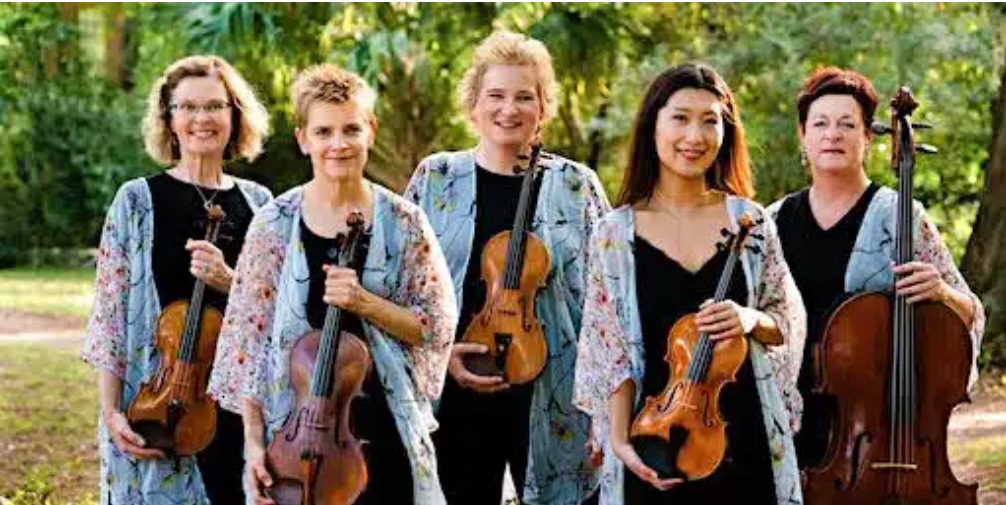 Five women stand outdoors, each holding a string instrument—violins, viola, and cello. They wear matching floral-patterned jackets over black outfits and smile at the camera, with greenery in the background.