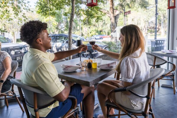 A man and a woman sit at an outdoor restaurant table, smiling and toasting with glasses of wine. Sunlight filters through trees, and table settings with plates and candles are visible.