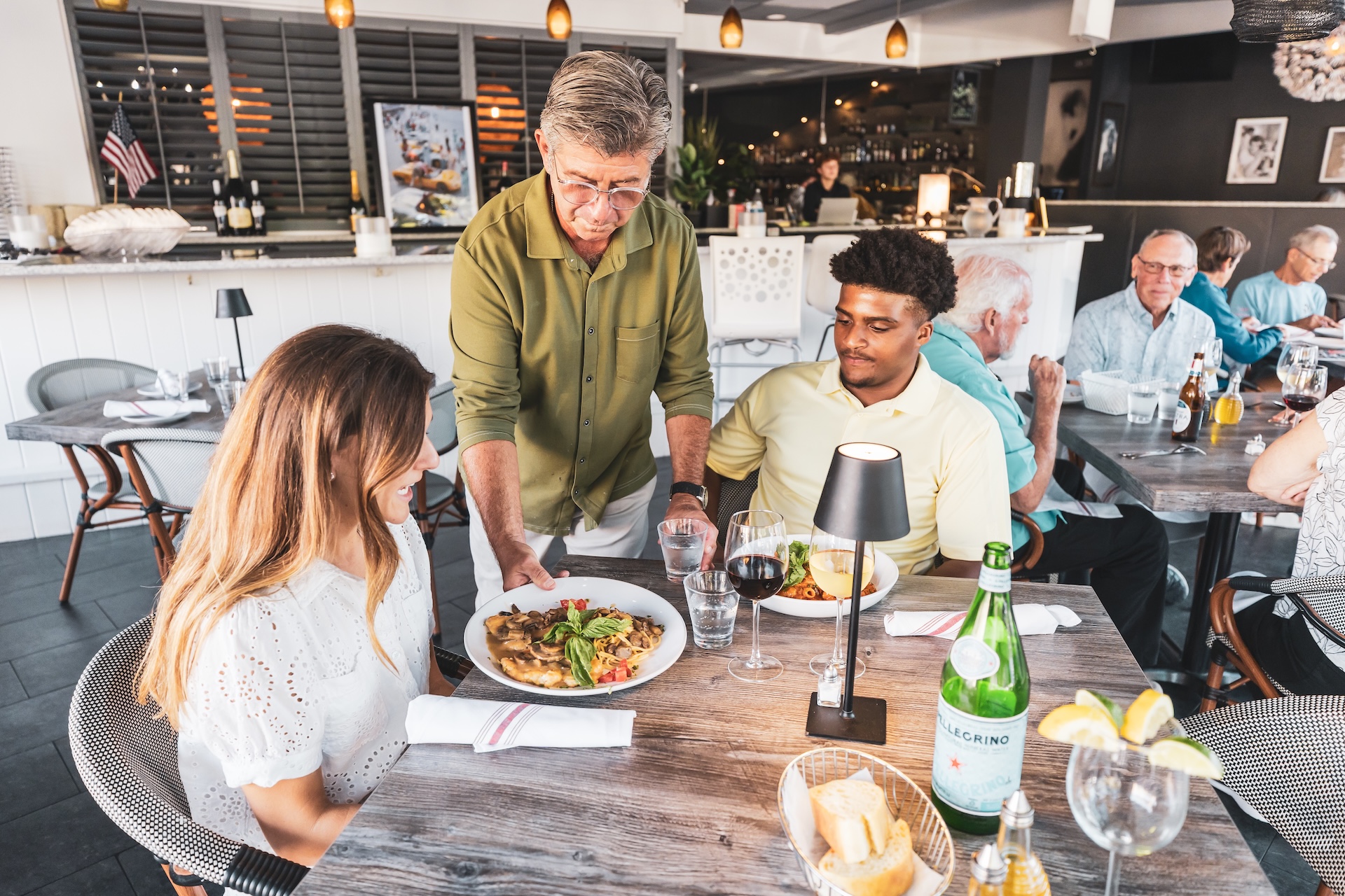 A waiter serves pasta to a woman at a restaurant table where she sits with a man. The table has water, wine, and bread. Other diners are seen in the background in a bright, modern dining area.