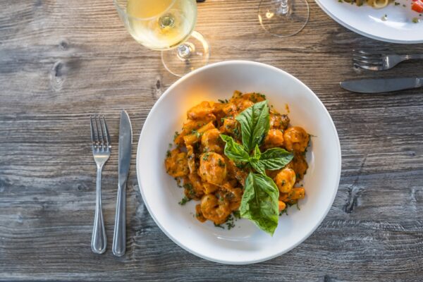 A white bowl filled with pasta in a creamy orange sauce, garnished with fresh basil leaves, placed on a wooden table next to a fork, knife, spoon, and a glass of white wine.