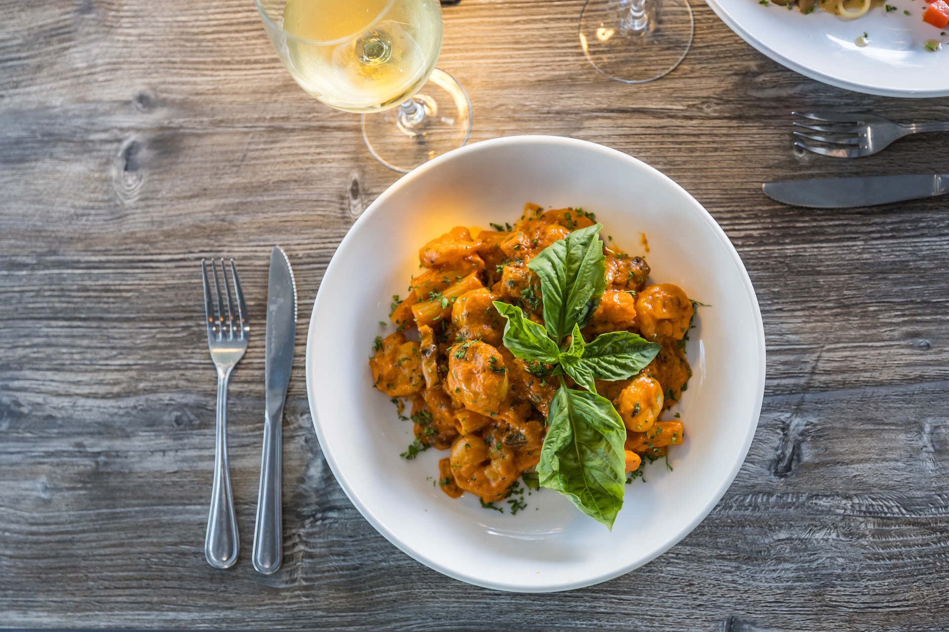 A white bowl filled with pasta in a creamy orange sauce, garnished with fresh basil leaves, placed on a wooden table next to a fork, knife, spoon, and a glass of white wine.