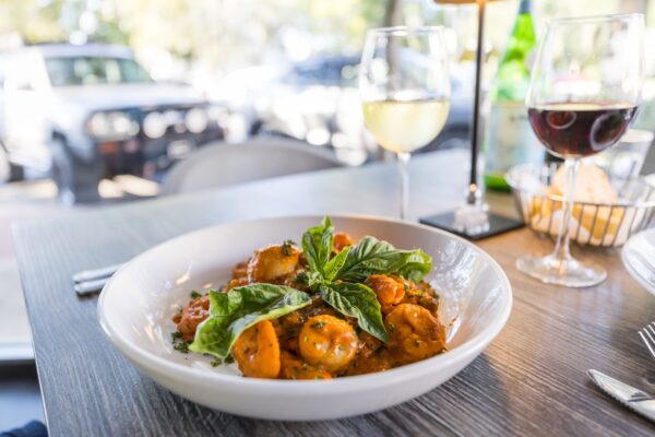 A white bowl of pasta with tomato sauce and fresh basil is on a table set with wine glasses, one filled with white wine and one with red, in a bright, casual dining setting near a window.