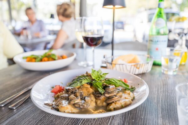 A plate of chicken with mushrooms and sauce garnished with basil sits on a wooden table with a glass of red wine, bread basket, bottled water, and another dish. People are blurred in the background at an outdoor restaurant.