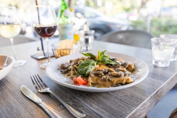 A plate of pasta with creamy mushroom sauce, garnished with fresh basil and tomato, sits on a wooden table set with cutlery, wine glasses, and water, with an outdoor setting visible in the background.