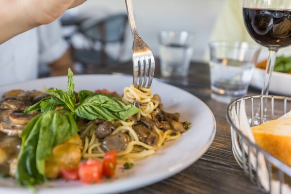 A close-up of a plate of spaghetti with mushrooms, fresh basil, and diced tomatoes. A hand is twirling spaghetti with a fork. A glass of red wine, water, and a basket of bread are on the wooden table.
