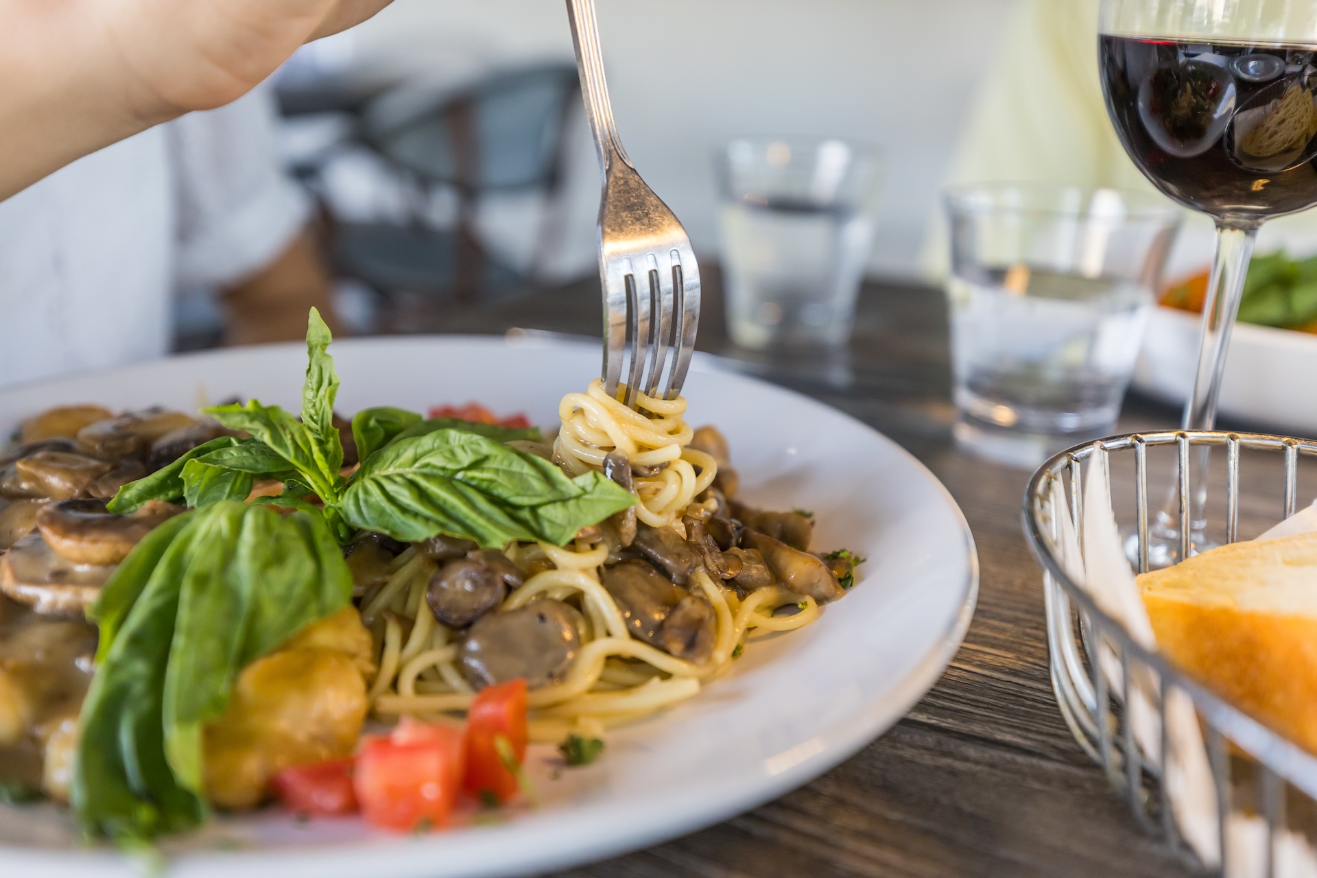 A close-up of a plate of spaghetti with mushrooms, fresh basil, and diced tomatoes. A hand is twirling spaghetti with a fork. A glass of red wine, water, and a basket of bread are on the wooden table.