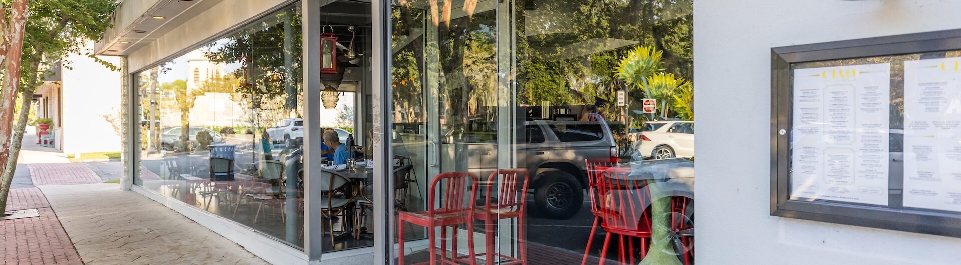 Street view of a modern restaurant with large windows, red chairs visible inside, a menu displayed outside, and trees reflecting on the glass facade. A brick sidewalk runs alongside the building.