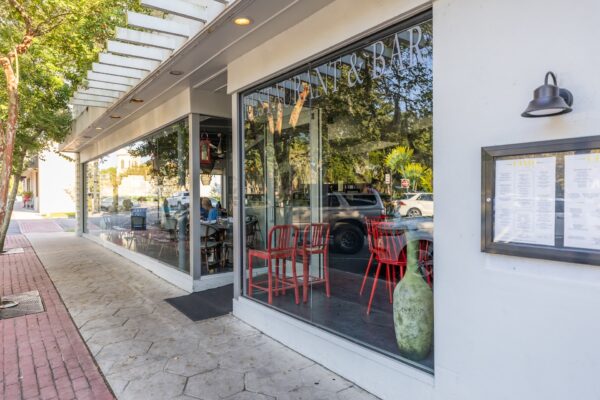 Street view of a modern restaurant with large windows, red chairs visible inside, a menu displayed outside, and trees reflecting on the glass facade. A brick sidewalk runs alongside the building.