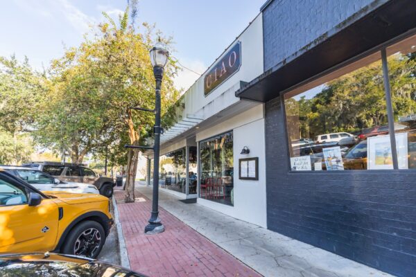 A sidewalk view of a street with parked cars, trees, a black lamppost, and storefronts, including a restaurant with a sign that reads GUIAO and outdoor seating visible through the windows.