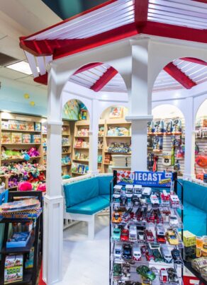 A colorful toy store with shelves full of stuffed animals, books, and various toys. A white gazebo with red trim and blue seats is in the center, and a display of die-cast toy cars is in the foreground.