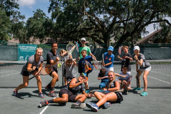 A group of people, both children and adults, pose happily with tennis rackets on an outdoor tennis court, with trees and a green fence in the background. Some are standing, others are sitting or kneeling.