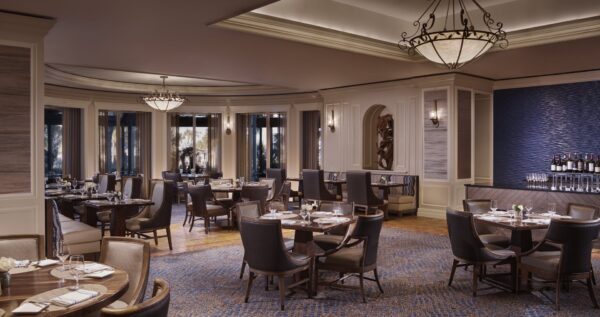 Elegant restaurant dining room with round tables set for guests, plush dark chairs, soft lighting from chandeliers, large windows, a blue accent wall, and a small bar area with wine bottles. The space is empty of people.