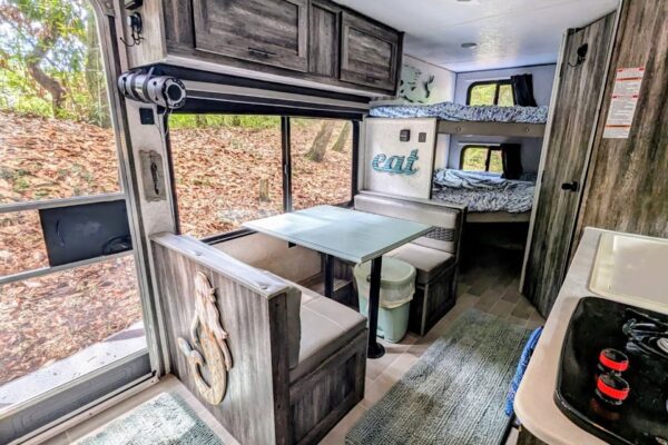 Interior of a cozy camper with a dining booth, table, bunk beds, and kitchen area. Sunlight comes through a large window, and nature can be seen outside. Nautical décor and modern finishes are visible throughout the space.