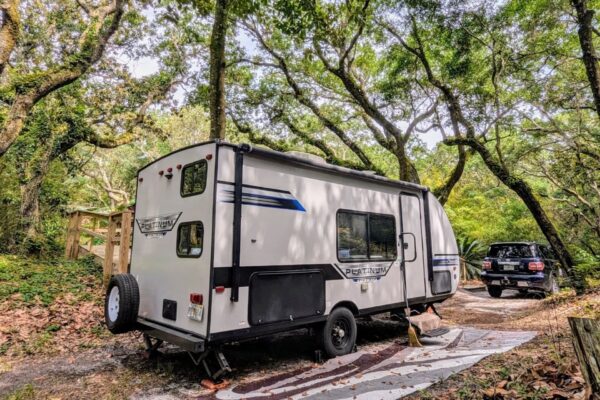 A white travel trailer is parked on a wooded campsite surrounded by tall, leafy trees. An SUV is parked nearby on a gravel path, and a striped outdoor mat lies beside the trailer. Sunlight filters through the green canopy.