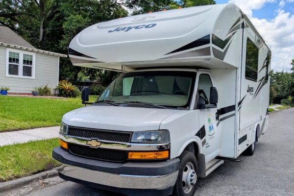 A white Jayco RV is parked on a suburban street next to a green lawn and a light-colored house. The RV features a Chevrolet front and bold black graphics. Trees and a partly cloudy sky are visible in the background.