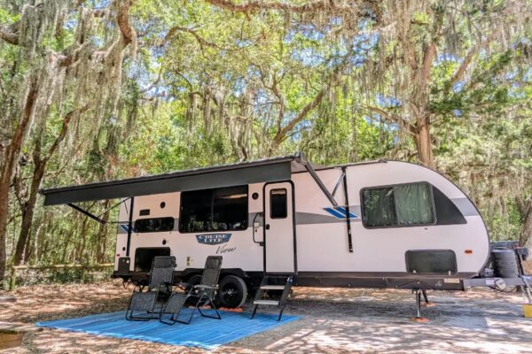 A white travel trailer with an extended black awning is parked in a shady, wooded campsite. Two reclining chairs and a blue mat are set up outside under hanging Spanish moss. Sunlight filters through the trees.