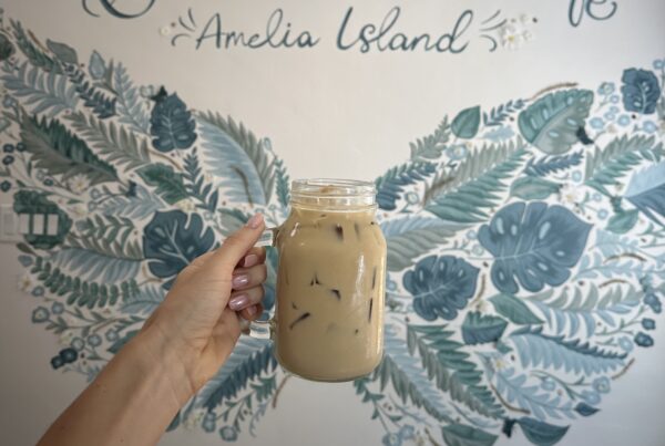 A hand holds a glass mason jar of iced coffee in front of a wall mural featuring the words Amelia Island surrounded by painted ferns and leaves.