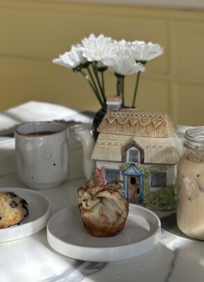 A blueberry scone and a pastry on plates next to a mug, a cottage-shaped teapot, and a glass of iced coffee on a marble table.