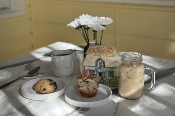 A blueberry scone and a pastry on plates next to a mug, a cottage-shaped teapot, and a glass of iced coffee on a marble table.