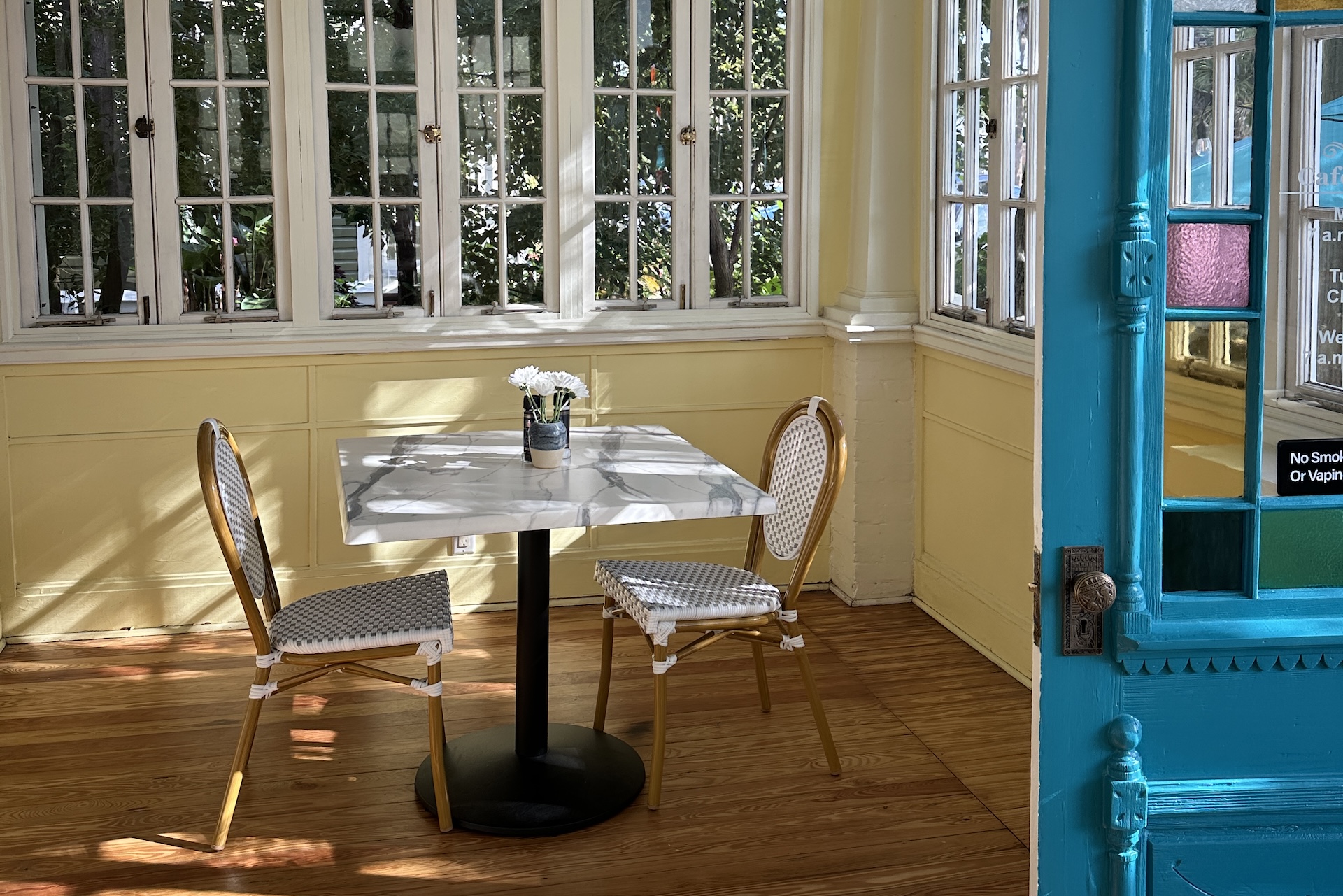 A marble-topped square table with two woven chairs and a small vase of white flowers, positioned in a sunny room with yellow walls and a bright blue door frame.
