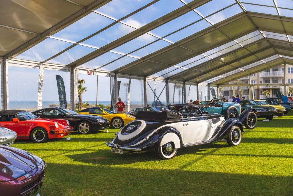 A classic black and white convertible car is displayed with other vintage and sports cars under a large transparent tent on a grassy area, with people walking around and buildings visible outside.