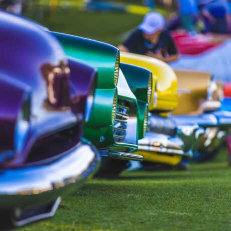 A row of colorful classic cars, including purple, green, yellow, and red, parked on grass at a car show, with a person in the background.
