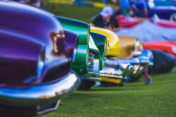 A row of colorful classic cars, including purple, green, yellow, and red, parked on grass at a car show, with a person in the background.