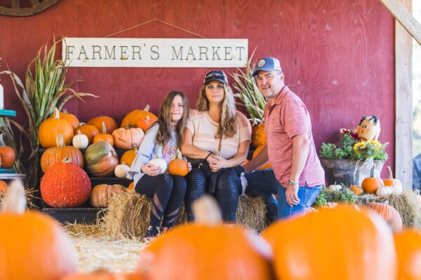 A family of three poses among pumpkins and hay bales in front of a red wall with a “Farmer’s Market” sign, at what appears to be a pumpkin patch or autumn market.