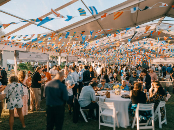 People gather under a tent decorated with colorful pennant banners for an outdoor event. Several tables with food are set up, and guests are eating, socializing, and enjoying the festive atmosphere.